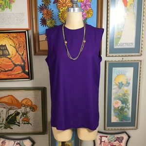 May include: A sleeveless purple dress with a gold and silver necklace. The dress is on a mannequin against a wall with framed artwork.