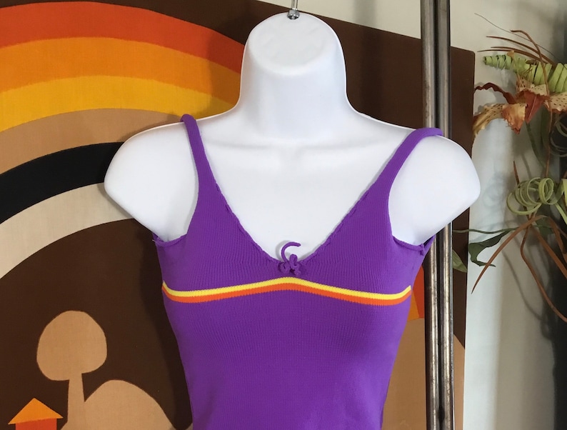 Pode incluir: Top roxo de malha canelada com uma faixa amarela e laranja no peito. O top tem um pequeno la&ccedil;o na gola.