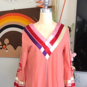 May include: A long-sleeved, peach-colored tunic with a V-neckline. The neckline is decorated with a multi-colored ribbon design in shades of pink, red, purple, and white.