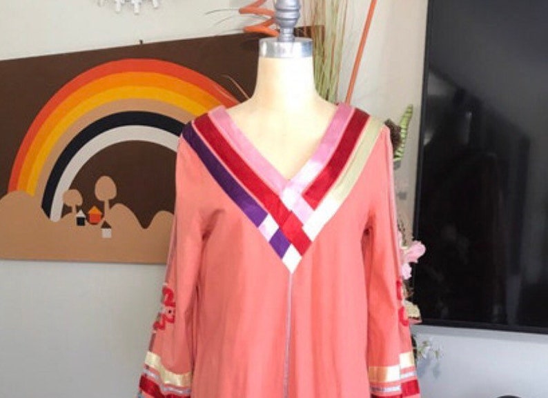 May include: A long-sleeved, peach-colored tunic with a V-neckline. The neckline is decorated with a multi-colored ribbon design in shades of pink, red, purple, and white.
