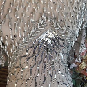 May include: A silver and white beaded top with a sequined design. The top is made of a sheer fabric and has a loose, flowing fit.