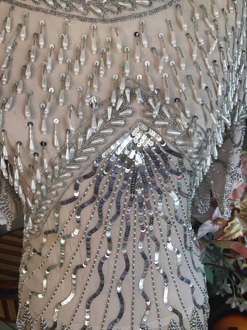 May include: A silver and white beaded top with a sequined design. The top is made of a sheer fabric and has a loose, flowing fit.