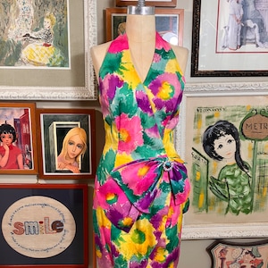 May include: A halter dress with a vibrant floral print in shades of pink, purple, green, and yellow. The dress has a bow detail at the waist and a fitted silhouette.