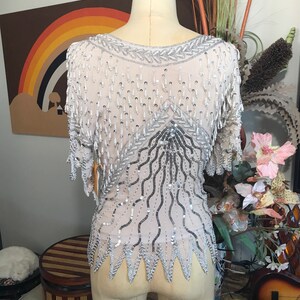 May include: A silver and white beaded top with a fringe design. The top is on a white dress form.