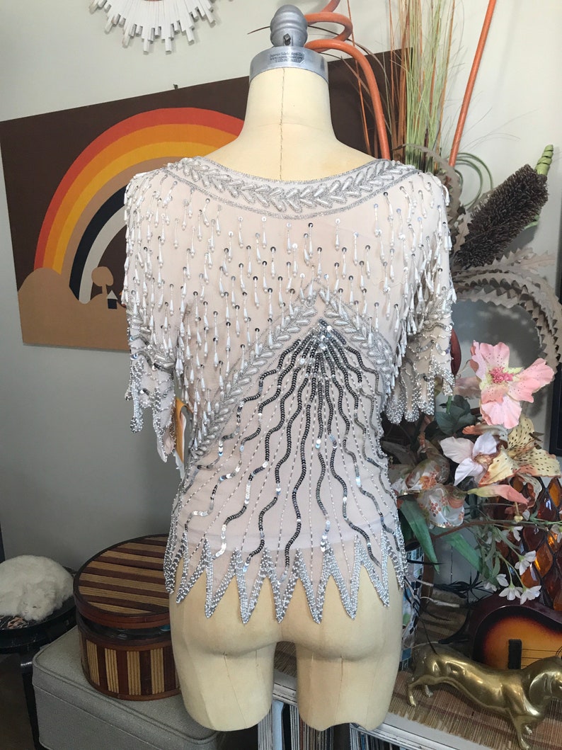 May include: A silver and white beaded top with a fringe design. The top is on a white dress form.