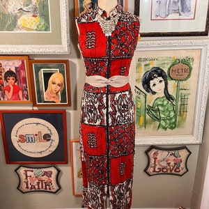 May include: A red and white sleeveless dress with a geometric pattern. The dress has a white belt and a beaded necklace. The dress is on a mannequin in front of a wall with artwork.