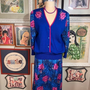 May include: A blue cardigan with a pink trim and floral embroidery on the sleeves, paired with a matching blue skirt with a floral print. The skirt is knee-length and has a fitted waist.