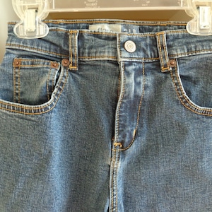 May include: A pair of blue denim jeans with a light wash. The jeans have a straight leg and a button fly. The waistband has a brown stitching detail. The jeans have a small pocket on the front right side.