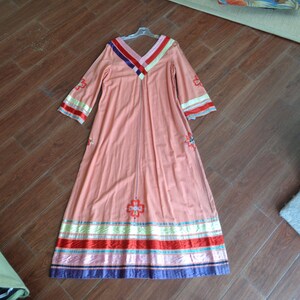 May include: A long, peach-colored maxi dress with embroidered floral details and colorful stripes around the hem. The dress has long sleeves and a V-neckline.