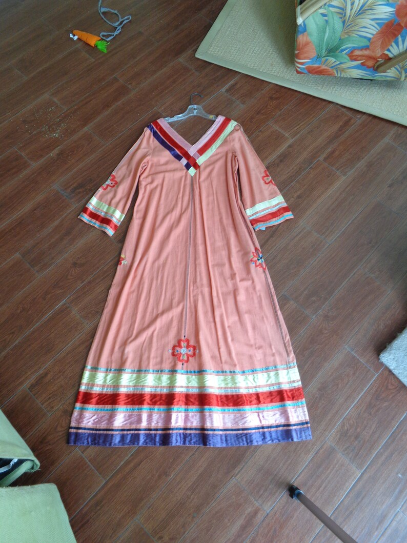 May include: A long, peach-colored maxi dress with embroidered floral details and colorful stripes around the hem. The dress has long sleeves and a V-neckline.