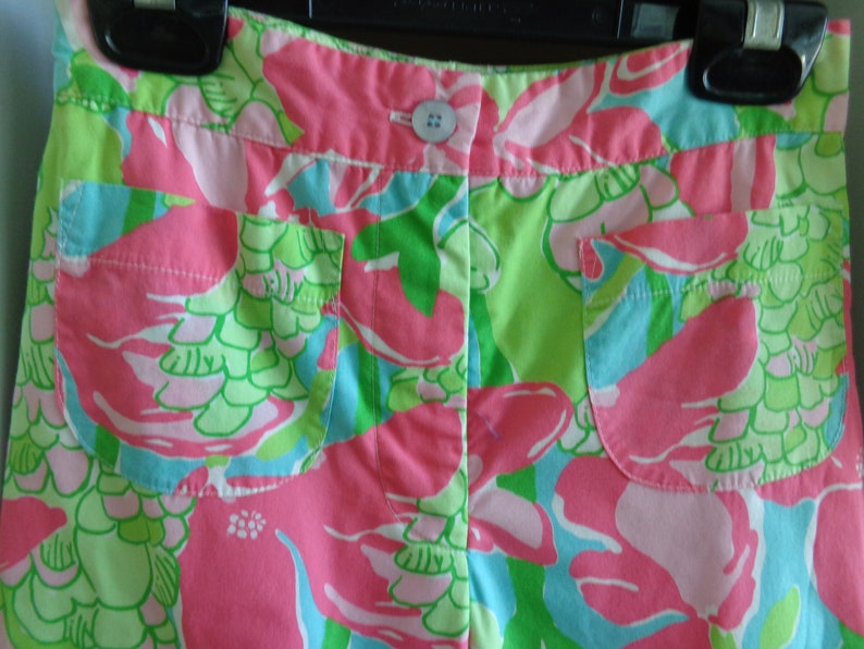 May include: Pink and green patterned shorts with two front pockets. The shorts have a button closure and a floral design.