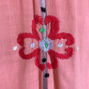 May include: A pink linen dress with a floral embroidery design. The flower is red with white and green accents. The dress has a purple and brown trim.
