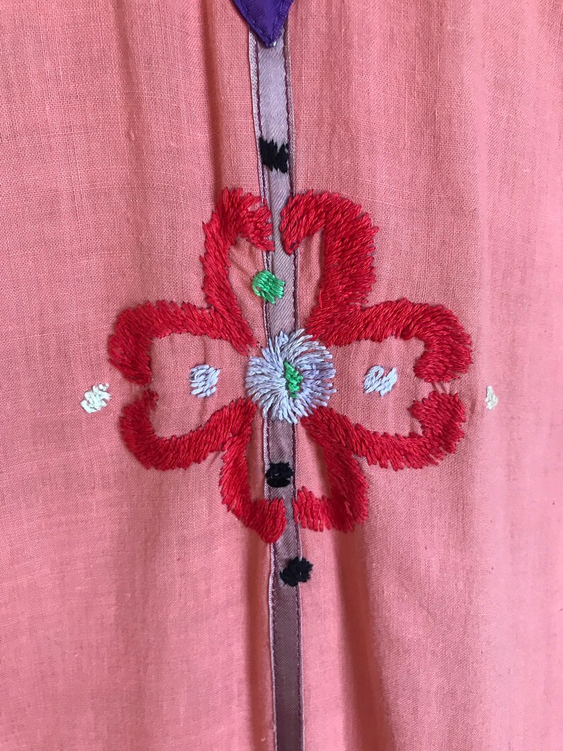 May include: A pink linen dress with a floral embroidery design. The flower is red with white and green accents. The dress has a purple and brown trim.