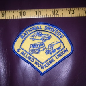 Könnte beinhalten: Ein gestickter Aufnäher in Gelb und Blau mit dem Text "National Drivers & Allied Workers Union". Der Aufnäher zeigt drei Fahrzeuge: ein Auto, einen Van und einen LKW.