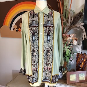 May include: A light green long-sleeved button-up shirt with a vintage-inspired print featuring ornate columns, floral motifs, and winged figures. The shirt has a classic collar and a straight hem.