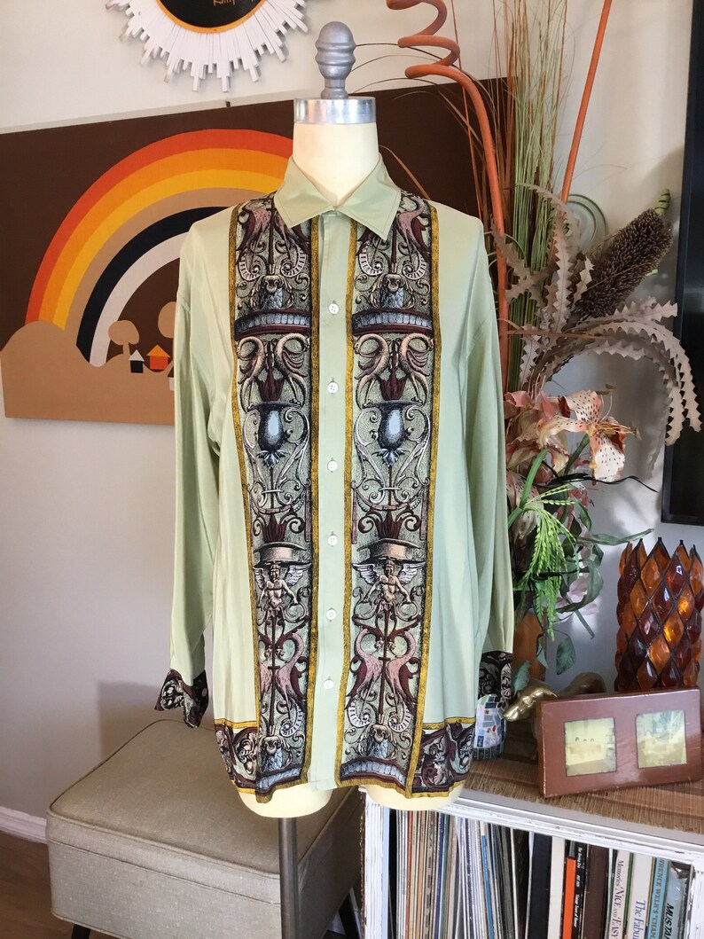 May include: A light green long-sleeved button-up shirt with a vintage-inspired print featuring ornate columns, floral motifs, and winged figures. The shirt has a classic collar and a straight hem.