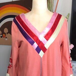 May include: A pink long-sleeved dress with a V-neckline and a colorful ribbon design on the neckline. The ribbon design features stripes of red, pink, purple, and white.