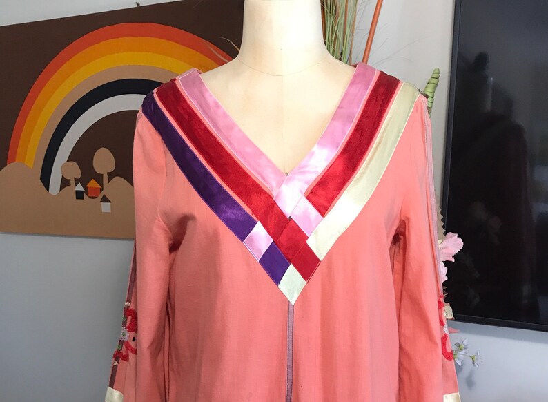 May include: A pink long-sleeved dress with a V-neckline and a colorful ribbon design on the neckline. The ribbon design features stripes of red, pink, purple, and white.