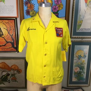 May include: A yellow short-sleeved bowling shirt with a pocket on the left side. The shirt has a black embroidered name "Lorene" on the left side of the chest and a red, white, and blue patch with the text "Brunswick 300" on the right side of the chest.