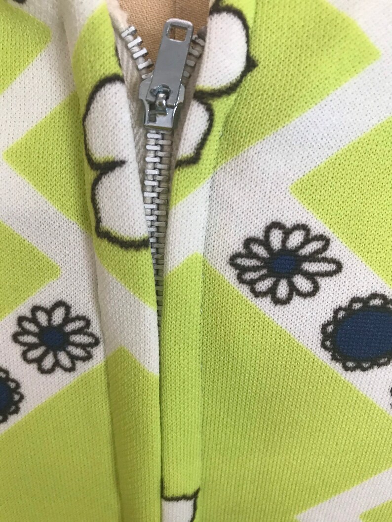 May include: A close-up of a zipper on a green and white patterned fabric. The fabric has a geometric pattern with white lines and black and white flowers.