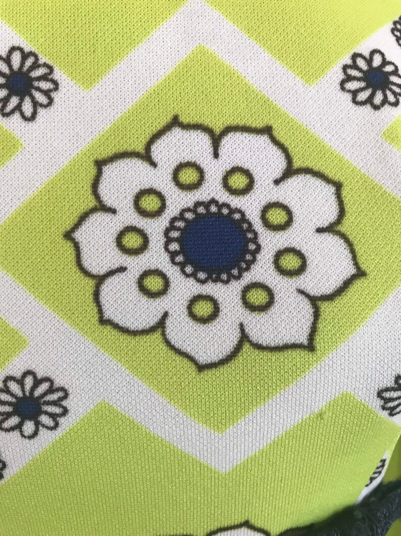 May include: A green fabric with a white and black geometric pattern. The pattern features a large white flower with a blue center and black outline.