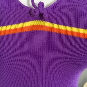 Pode incluir: Camisola de malha roxa com uma faixa amarela, laranja e vermelha no peito. A camisola tem um la&ccedil;o no decote.