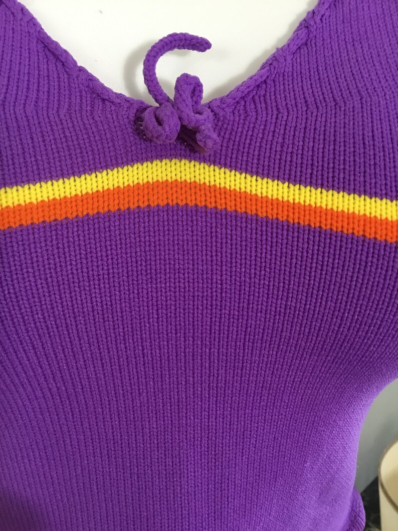 Pode incluir: Camisola de malha roxa com uma faixa amarela, laranja e vermelha no peito. A camisola tem um la&ccedil;o no decote.