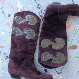 May include: A pair of brown suede knee-high boots with green snakeskin accents. The boots have a pointed toe and a low heel.