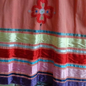 May include: A pink skirt with a colorful embroidered flower design and a multi-colored striped trim. The trim includes red, pink, blue, and green ribbons.