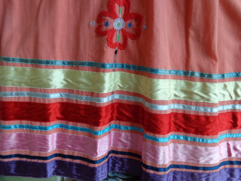 May include: A pink skirt with a colorful embroidered flower design and a multi-colored striped trim. The trim includes red, pink, blue, and green ribbons.
