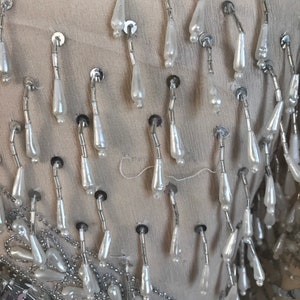 May include: Close-up of a garment with silver and white beaded fringe. The fringe is made of small, teardrop-shaped beads that are strung together on silver wire.