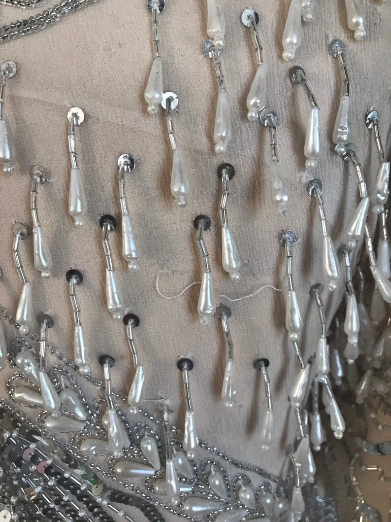May include: Close-up of a garment with silver and white beaded fringe. The fringe is made of small, teardrop-shaped beads that are strung together on silver wire.