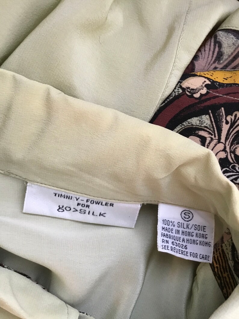 May include: A green silk blouse with a brown, black, and gold patterned design. The tag reads "Timniy-Fowler for 100% Silk" and "100% Silk/Soie Made in Hong Kong Fabrique a Hong Kong RN 63026 See Reverse for Care."