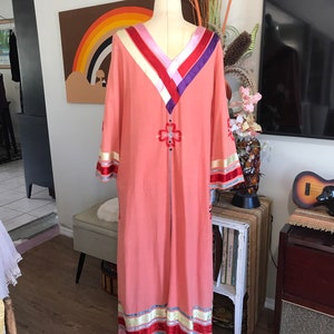 May include: A long, peach-colored maxi dress with a V-neckline and long sleeves. The dress has a colorful embroidered design on the front and is trimmed with colorful ribbon.