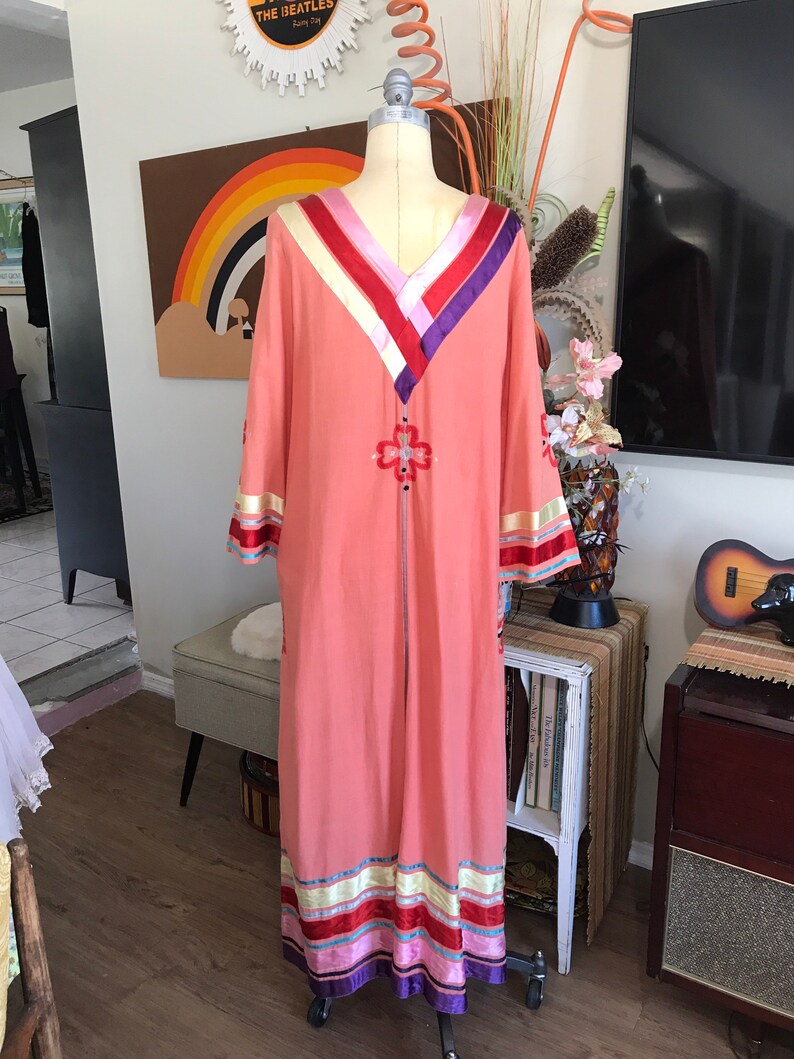 May include: A long, peach-colored maxi dress with a V-neckline and long sleeves. The dress has a colorful embroidered design on the front and is trimmed with colorful ribbon.