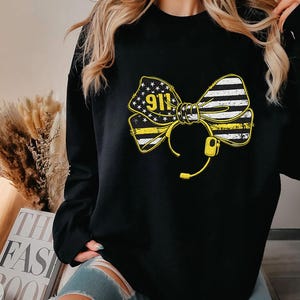 Puede incluir: Sudadera negra con un diseño de lazo amarillo y blanco, el número "911" y un gráfico de auriculares. El lazo incorpora elementos de la bandera estadounidense, con estrellas y rayas. La sudadera es de estilo casual.