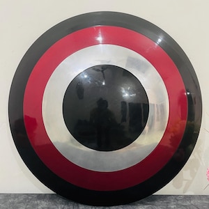 May include: A round shield with a black, red, white, and silver color scheme. The shield has a central black circle, surrounded by a silver ring, a red ring, and a black outer rim. The shield is displayed against a neutral background.