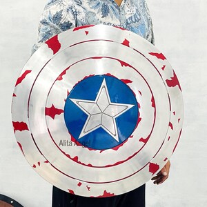 May include: A silver-colored shield with red accents and a blue circle featuring a white star in the center. The shield has a distressed appearance, with visible wear and tear. The shield is a replica of Captain America's shield.