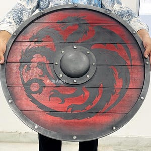 May include: A circular wooden shield featuring a red and black dragon motif. The shield has a dark grey metal rim and a central boss. The dragon emblem is displayed on the wooden surface, with a distressed finish.
