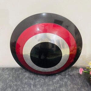 May include: A round shield with a black outer ring, a red middle ring, a silver inner ring, and a black center. The shield is a replica of a superhero's shield.