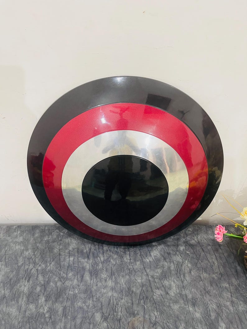 May include: A round shield with a black outer ring, a red middle ring, a silver inner ring, and a black center. The shield is a replica of a superhero's shield.