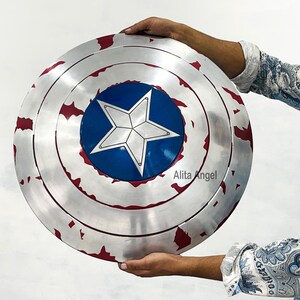 May include: A silver and red shield with a blue center and a white star. The shield has a distressed, battle-worn appearance. The shield is round and has concentric circles. The shield is being held.