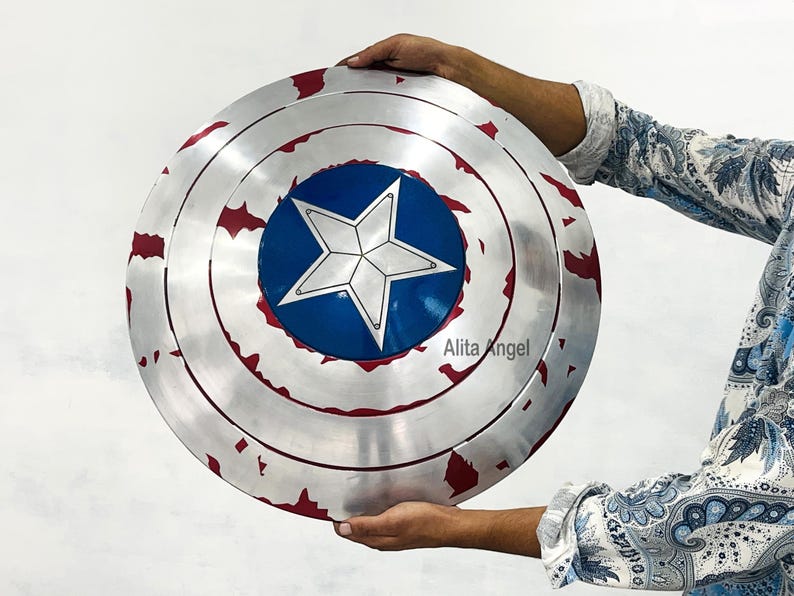May include: A silver and red shield with a blue center and a white star. The shield has a distressed, battle-worn appearance. The shield is round and has concentric circles. The shield is being held.