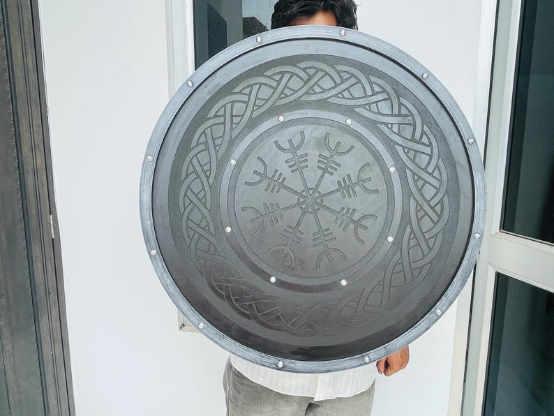 May include: A large, round shield with a dark gray finish and a silver trim. The shield features an intricate Celtic knot design around the edge and a central symbol. Small silver rivets are evenly spaced around the shield's perimeter.