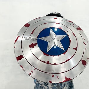 May include: A silver-colored, round shield with a blue center featuring a white star. The shield has red accents around the edges, and a distressed, battle-worn appearance. The shield is a prop or costume piece.