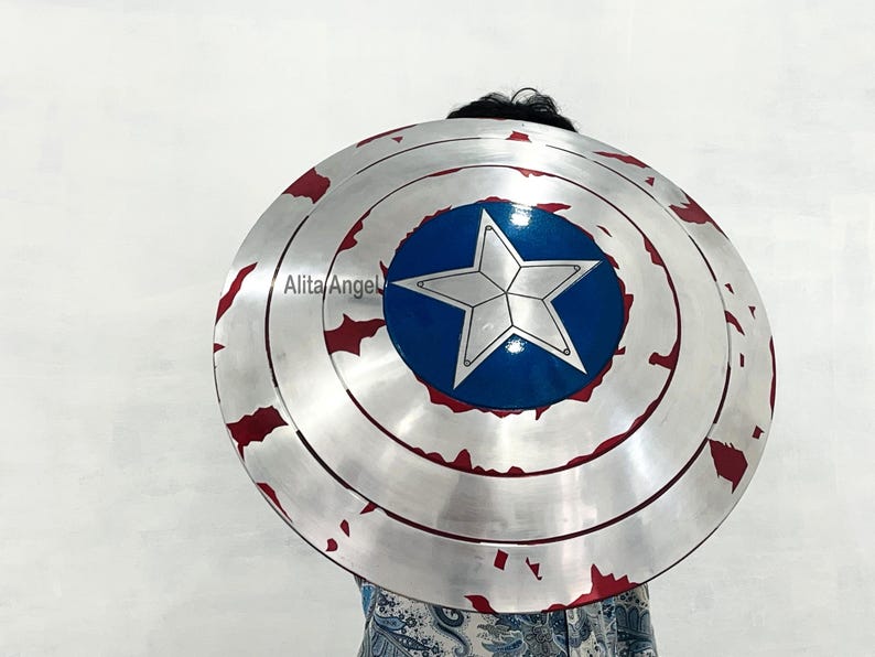 May include: A silver-colored, round shield with a blue center featuring a white star. The shield has red accents around the edges, and a distressed, battle-worn appearance. The shield is a prop or costume piece.