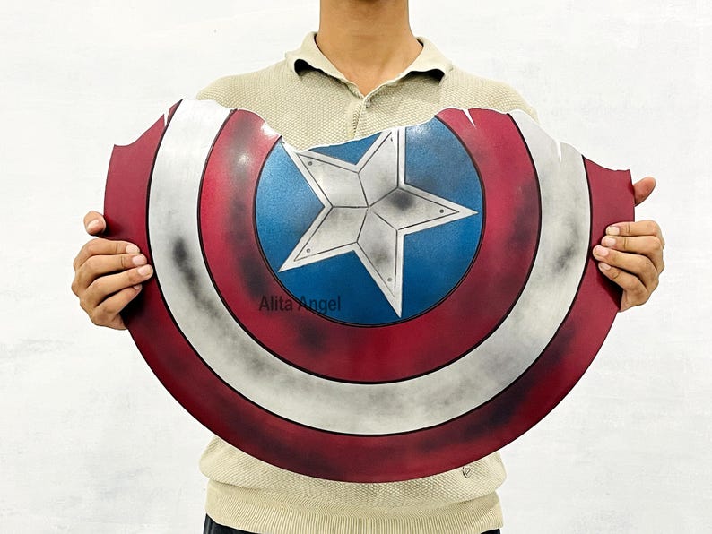 K&ouml;nnte beinhalten: Ein abgenutzter Captain America-Schild mit einem rot-wei&szlig;-blauen Farbschema und einem zentralen Sternemblem. Der Schild hat ein verwittertes Aussehen mit sichtbaren Abnutzungserscheinungen. Es ist eine Requisite oder ein Sammlerst&uuml;ck.