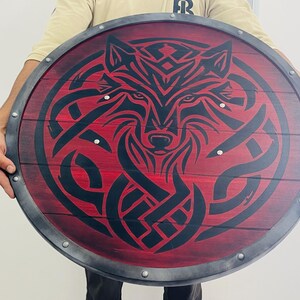 May include: Round wooden shield with a red background and a black wolf design. The shield has a metal rim and is held by a person. The wolf design is intricate and detailed, with a focus on the animal's face.