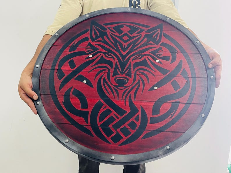 May include: Round wooden shield with a red background and a black wolf design. The shield has a metal rim and is held by a person. The wolf design is intricate and detailed, with a focus on the animal's face.