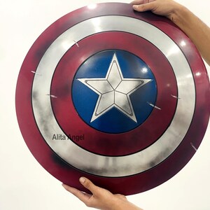 May include: A round shield with a red, white, and blue color scheme. The shield features a white star on a blue center, surrounded by concentric silver and red bands. The shield is held by hands.